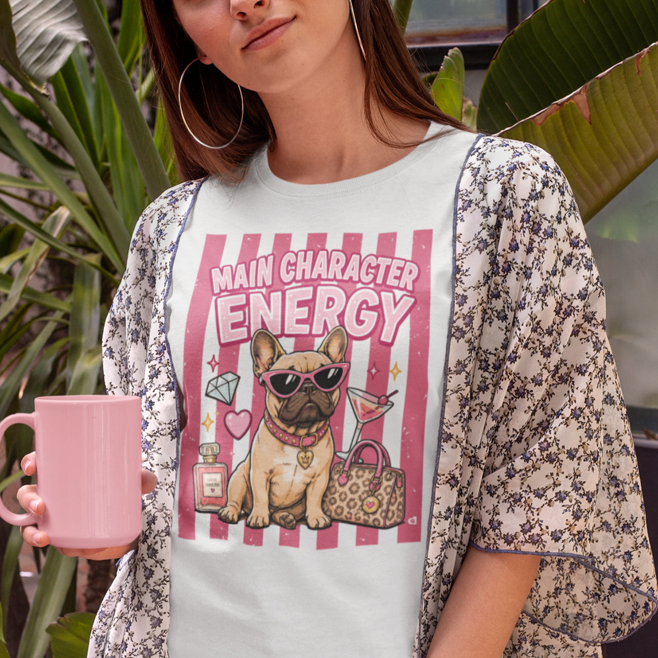 A woman wearing a white graphic T-shirt with a pink and red striped design featuring a French bulldog and the text 'MAIN CHARACTER ENERGY,' layered under a floral kimono-style cover-up. She is holding a pink mug in front of a tropical plant background.