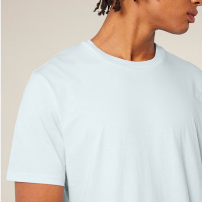A close-up of a person wearing a Stanley Stella short-sleeve crew-neck t-shirt in a very light icy blue colour, set against a solid warm beige background.